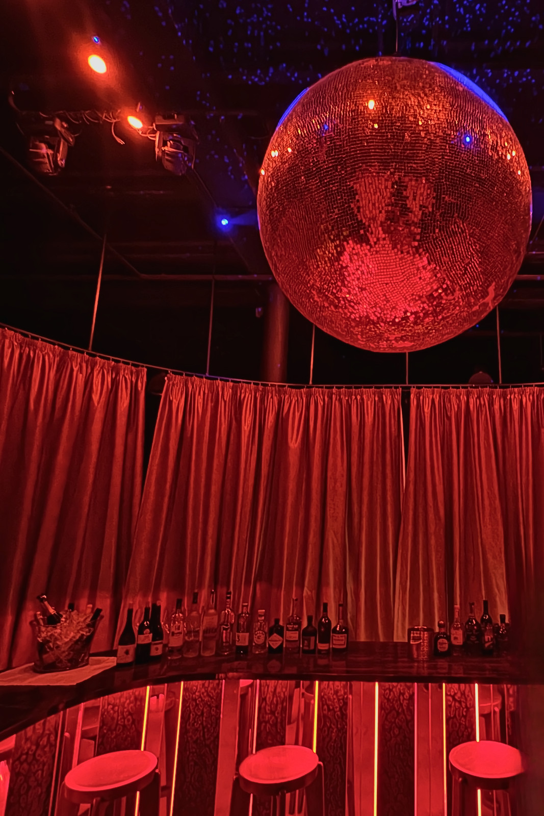 A large mirror ball under a starry ceiling above a curved bar ringed by red curtains and red-lit stools.