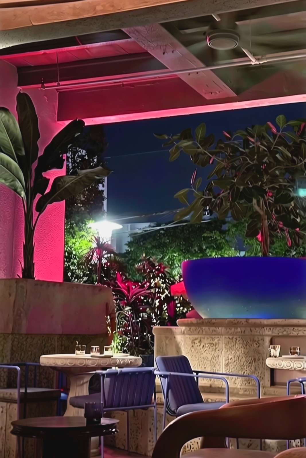 A covered patio lit pink at night, with a ceiling fan, a ficus in a large blue bowl planter, stone tables and blue chairs.