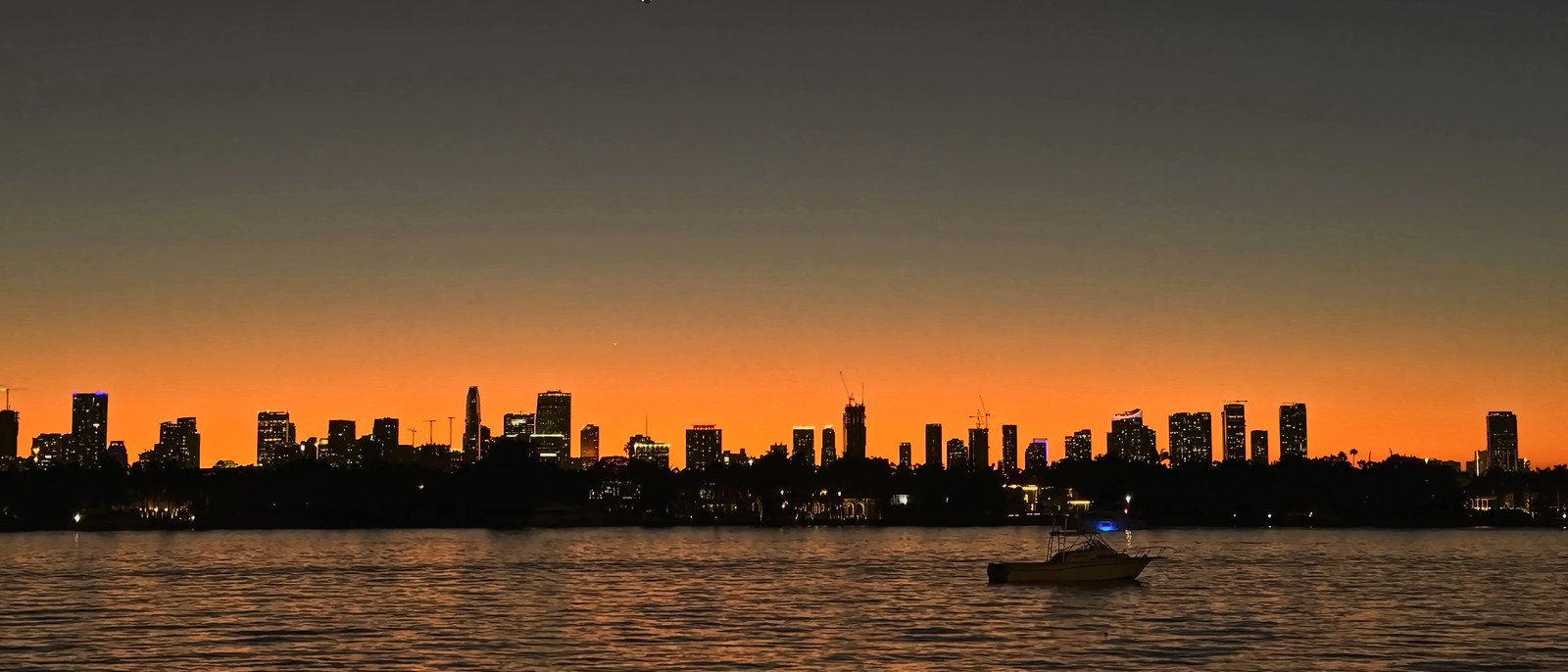 The Miami skyline silhouetted against an orange horizon across Biscayne Bay at dusk, with a small boat on the water.