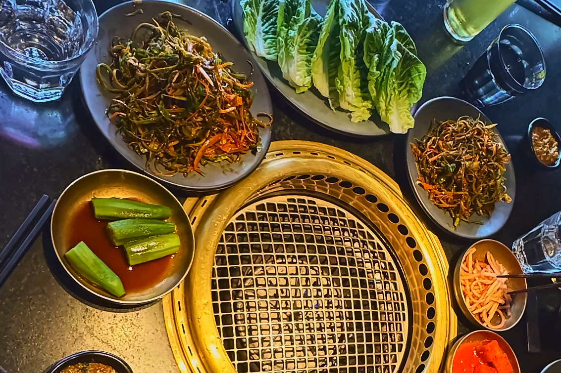 A brass tabletop grill surrounded by banchan, lettuce wraps and sauces on a dark table, seen from above.