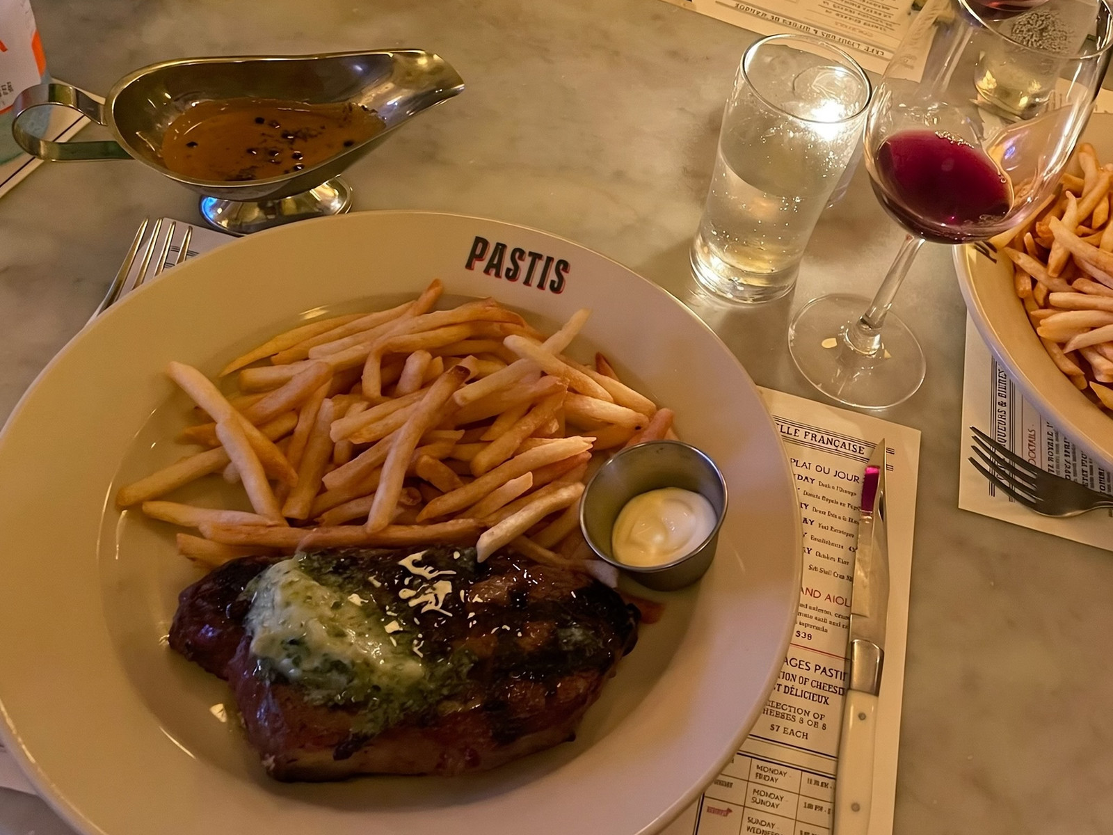 Steak frites with herb butter on a plate printed with PASTIS, with a boat of peppercorn sauce, aioli, the menu and a glass of red wine on a marble table.