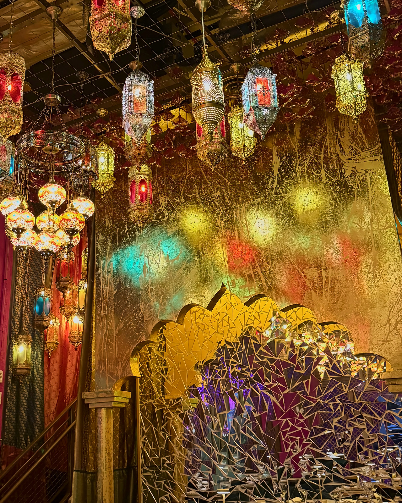 Colored metal lanterns hanging over a gold wall and a scalloped arch covered in mirror mosaic in an Indian restaurant dining room.