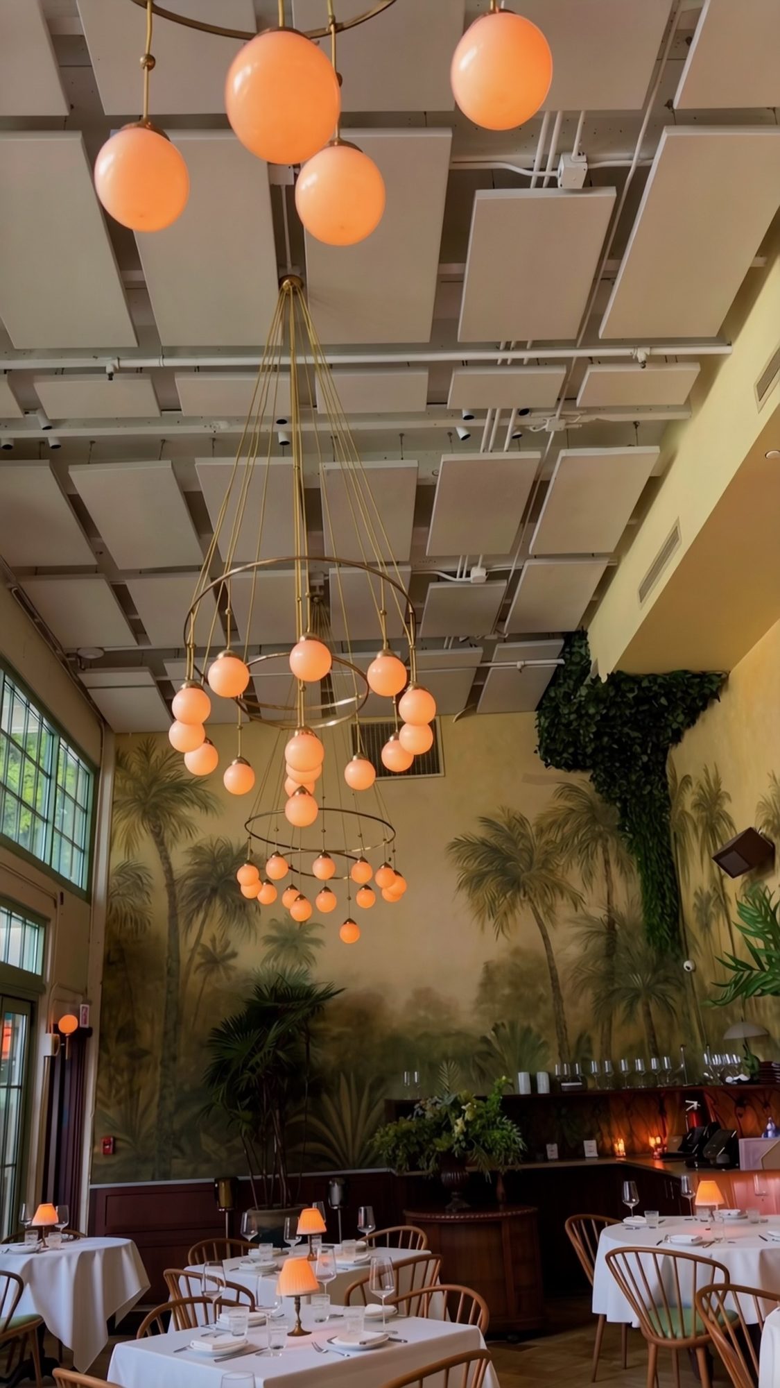 Globe chandeliers hanging from a high ceiling over a dining room with a painted palm mural and tables set with white cloths.