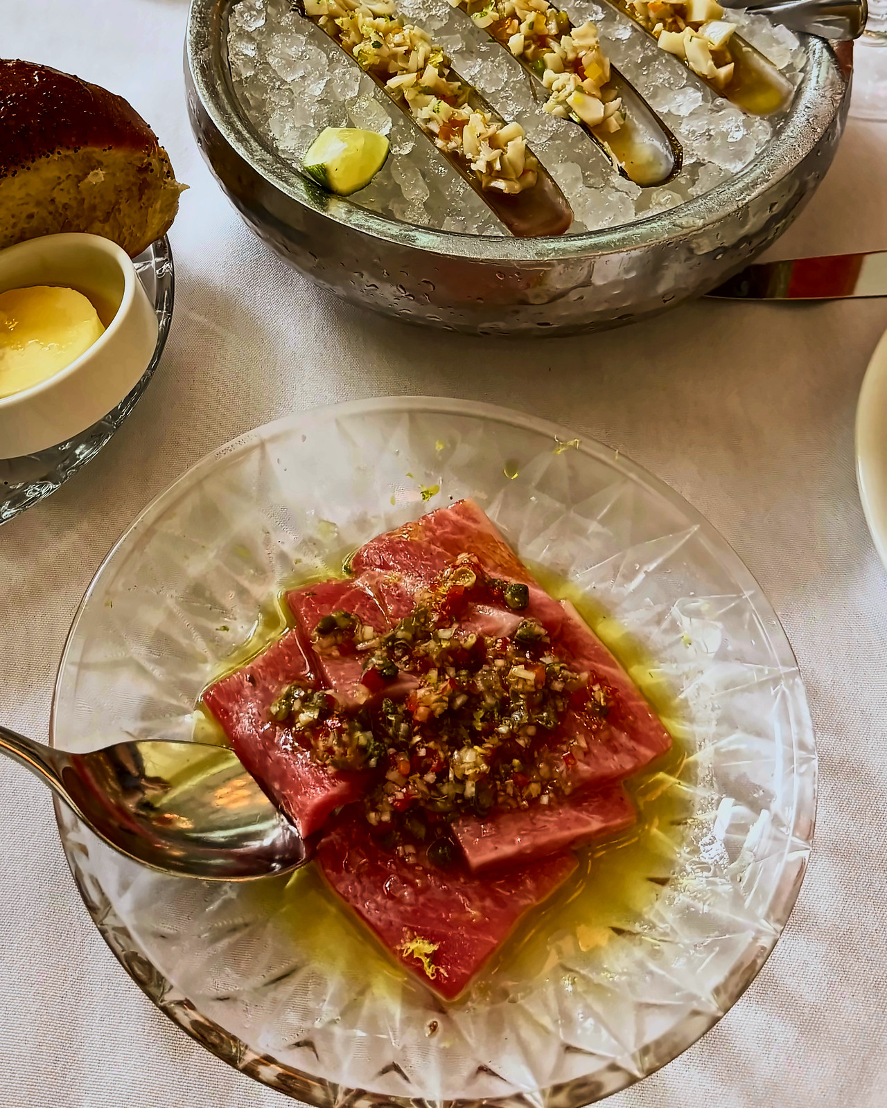 Razor clams on crushed ice and tuna crudo in a cut-glass dish on a white tablecloth.