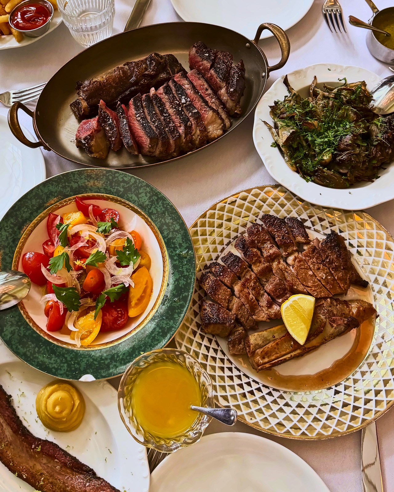 Sliced steak in an oval pan with brass handles, a grilled ribeye with a lemon wedge, a tomato and onion salad and sautéed mushrooms on a white tablecloth.
