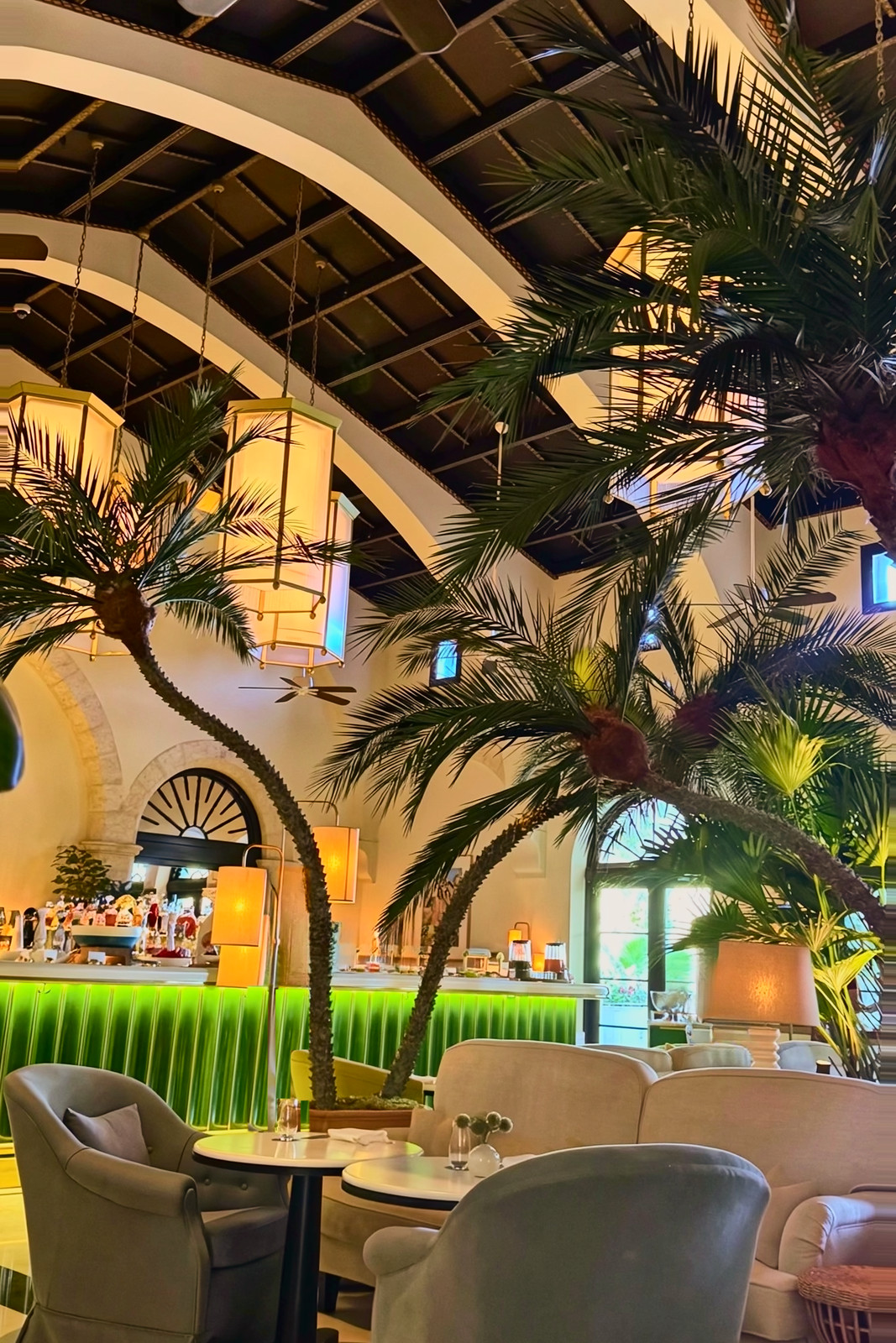 A green-lit bar under arched ceiling beams, with hanging lanterns, potted palms, lamps and armchairs in the foreground.