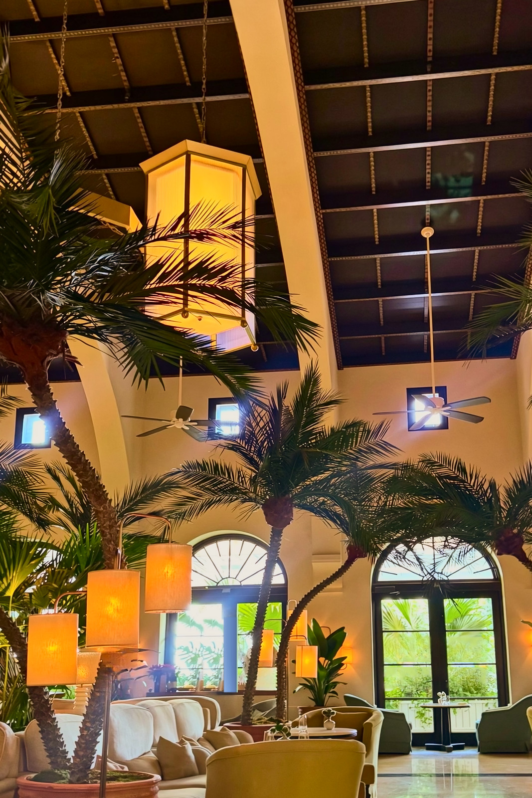 A double-height lounge with potted palms, hanging lanterns, arched garden doors and armchairs under a dark coffered ceiling.
